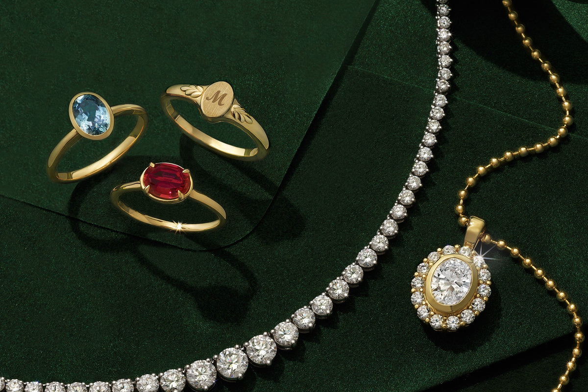 A jewelry set arranged on a dark green satin background, including a gold necklace with a diamond-studded pendant, a matching diamond tennis necklace, a pair of diamond hoop earrings, and three gold rings with blue, red, and engraved stones.